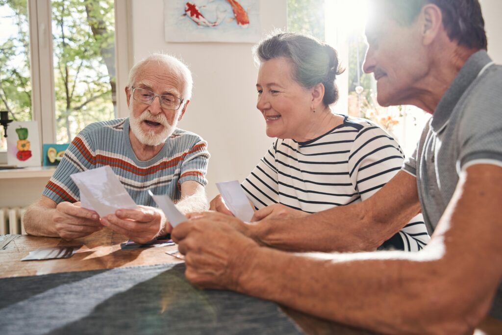 Three older adults sit at a sunny window, smiling as they look at photographs or cards together—perhaps discussing memories with their Authorized Representatives or Rep Payees—enjoying a relaxed and cheerful moment indoors.