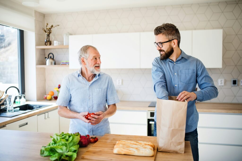 Two men in a bright kitchen prepare food together, enjoying the moment. As fresh bread and leafy greens sit on the counter, their teamwork embodies the supportive bond often seen in guardianship despite guardians’ limitations.