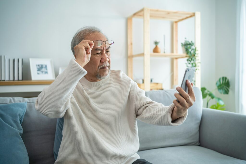 An older man sitting on a sofa holds up a smartphone and raises his glasses, looking closely at the screen—perhaps reading about Social Security scams. The background shows shelves with decorative items and plants in a bright living room.
