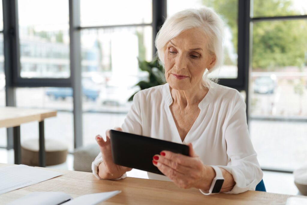 An older woman with gray hair, wearing a white blouse and a smartwatch, sits at a desk and looks at a tablet. She appears focused, perhaps reading about new wave scams like the grandma scam. Large windows and papers surround her.
