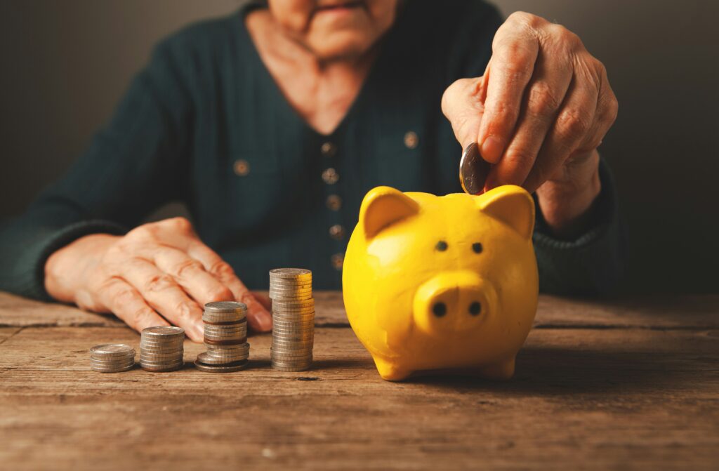 An older adult places a coin into a yellow piggy bank on a wooden table, with several stacks of coins beside it—a reminder for older adults to recognize scams and protect their savings from investment scams.