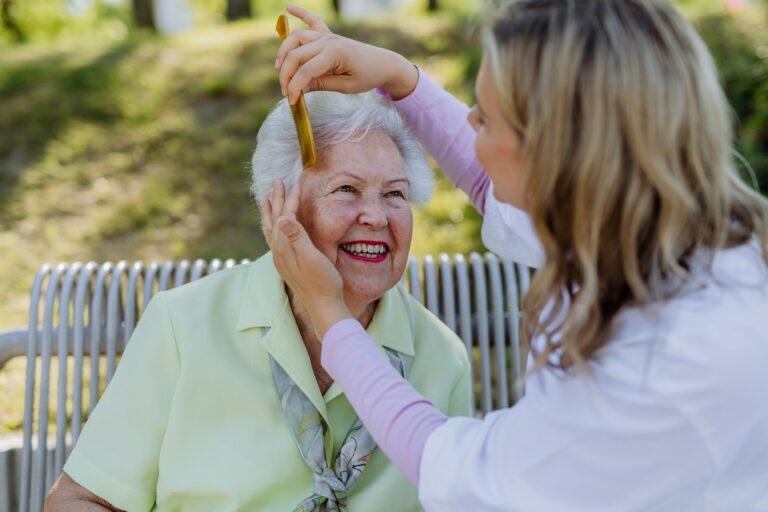 A young woman gently combs the hair of a smiling elderly woman with white hair, who is sitting on an outdoor bench and wearing a pale yellow blouse—capturing a warm, cheerful moment that reflects the caring spirit behind Minnesota Medical Assistance.