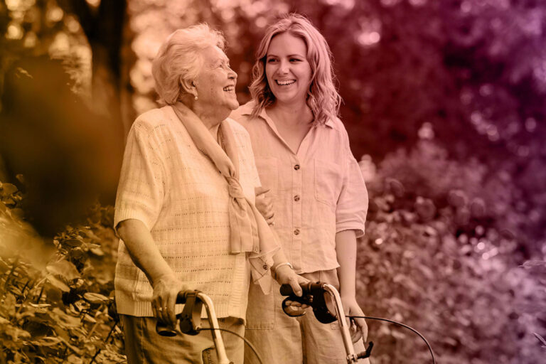 An elderly woman with a walker and a younger woman stand outdoors, smiling and laughing together. With lush greenery behind them and a warm, colorful gradient overlay, the image captures the spirit of connection—reflecting Everbright Legacy Law’s caring support.