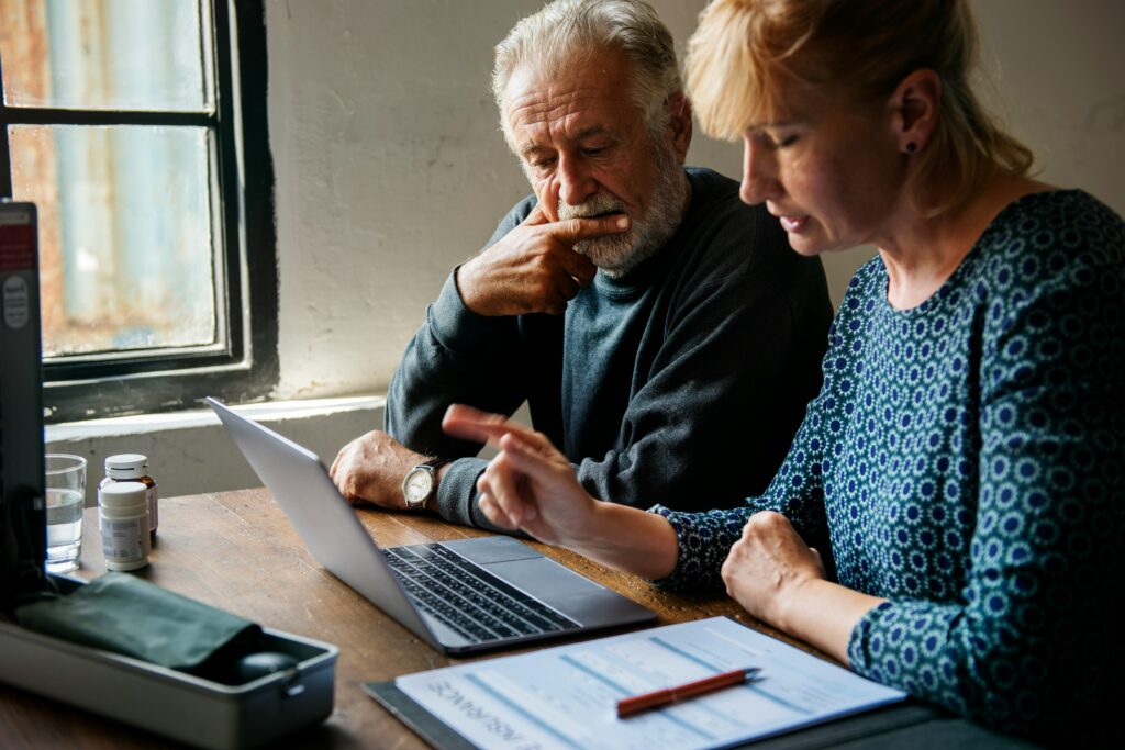 An older man and a woman sit at a table in Minnesota, looking at a laptop screen. The woman points at the screen, and there are documents, a pen, medicine bottles, and information about long-term care on the table near them.