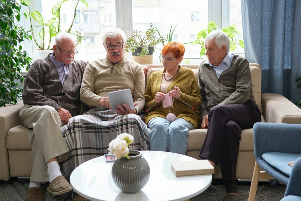 Four elderly people sit on a sofa; one man holds a tablet, another watches, while a woman knits and another man looks on. Surrounded by plants and with a round table featuring a vase and book in front, they gather together—sharing memories and finding ways to safeguard your legacy in Minnesota.