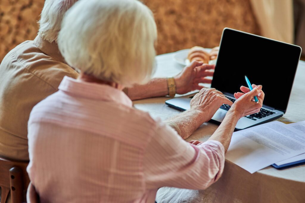 Two older adults sit at a table using a laptop, with one person pointing at the screen with a pen. Papers and a plate of pastries are on the table, suggesting they are thoughtfully reviewing their estate plan to ensure their wishes are honored.