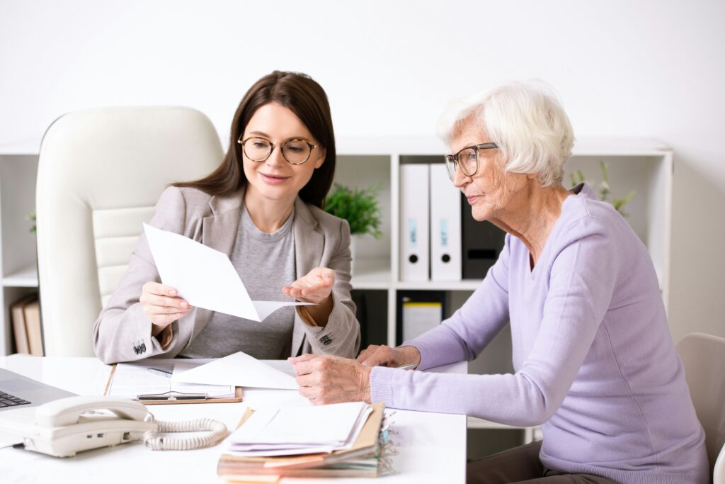 A younger woman in business attire shows a document to an older woman with white hair and glasses, both sitting at a desk with papers and office supplies. The setting reflects the trusted guidance of a reliable Minnesota elder law attorney assisting her client with important legal documents.