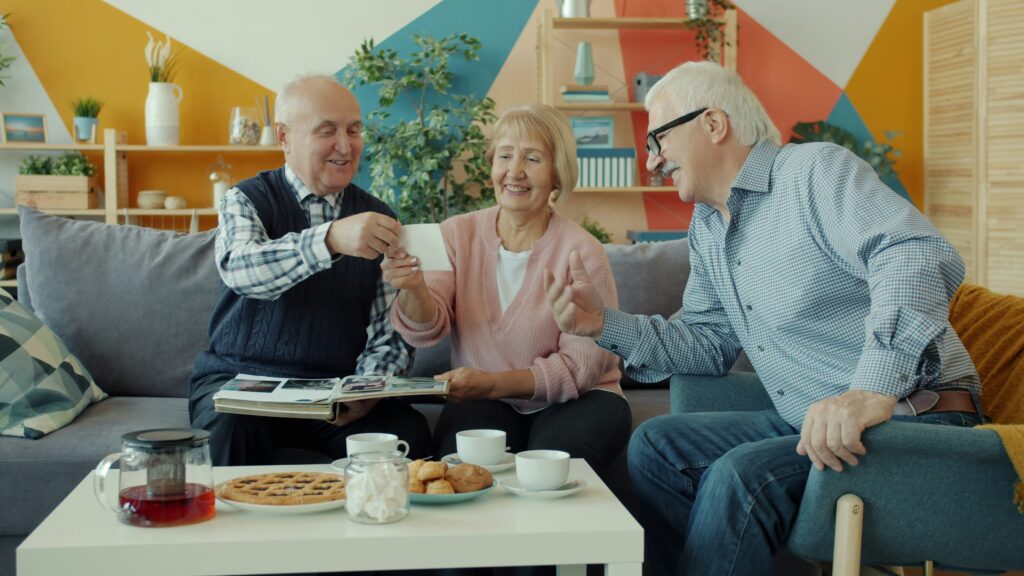 Three seniors sit on a couch, smiling and laughing together over a photo album. A table in front of them holds pastries, cookies, and cups of tea or coffee in a brightly decorated living room with plants and shelves—a warm setting that reminds you how important it is to safeguard your legacy Minnesota style: with cherished memories shared among friends.
