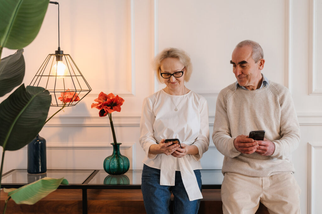 Two older adults sit together indoors, each looking at their smartphone. Behind them are red flowers in vases on a table, a hanging lamp, and white paneled walls.