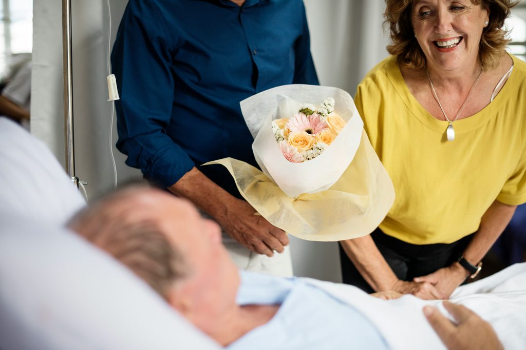 A woman and a man stand by a hospital bed, smiling and holding a bouquet of flowers, visiting a patient who is lying down—demonstrating the support and compassion often shown by a health care agent in Minnesota.
