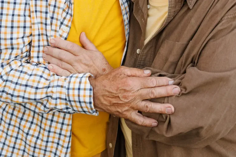 Two people stand close together, each with a hand gently resting on the others arm. Both are wearing casual shirts—one plaid and one brown. The image focuses on their hands and torsos, not showing their faces.