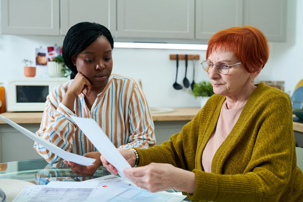 Two women sit at a table in a kitchen, reviewing papers together. One woman—possibly acting as a personal representative in Minnesota—has short red hair and glasses; the other has braided hair and wears a striped shirt. The table is covered with documents.