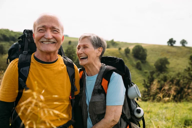 Two older adults with backpacks smile and laugh together while hiking in a grassy, hilly landscape on a sunny day.