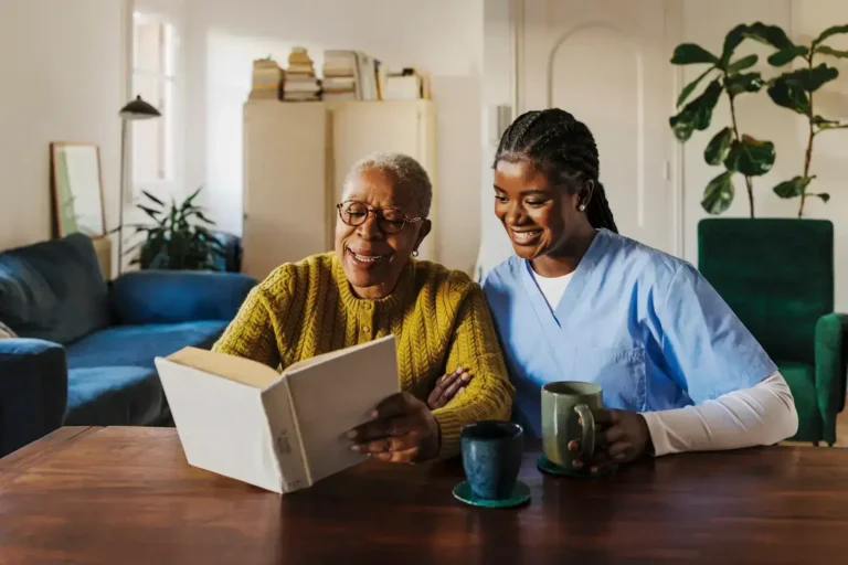 An elderly person and a caregiver sit at a table, smiling as they look at a book together. Both have mugs in front of them in a cozy, well-lit living room with plants and books in the background.