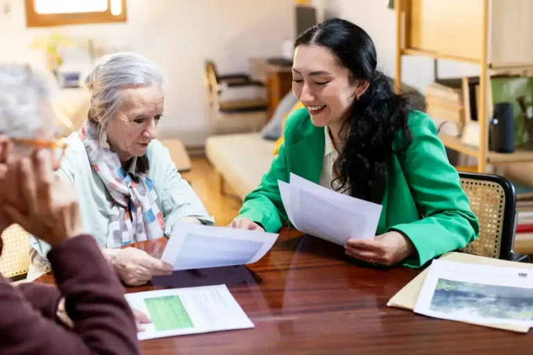 Three women sit at a wooden table reviewing documents together. One woman wears a green blazer and smiles, while another, with gray hair and a scarf, looks at her papers. The setting appears to be a cozy, well-lit room.