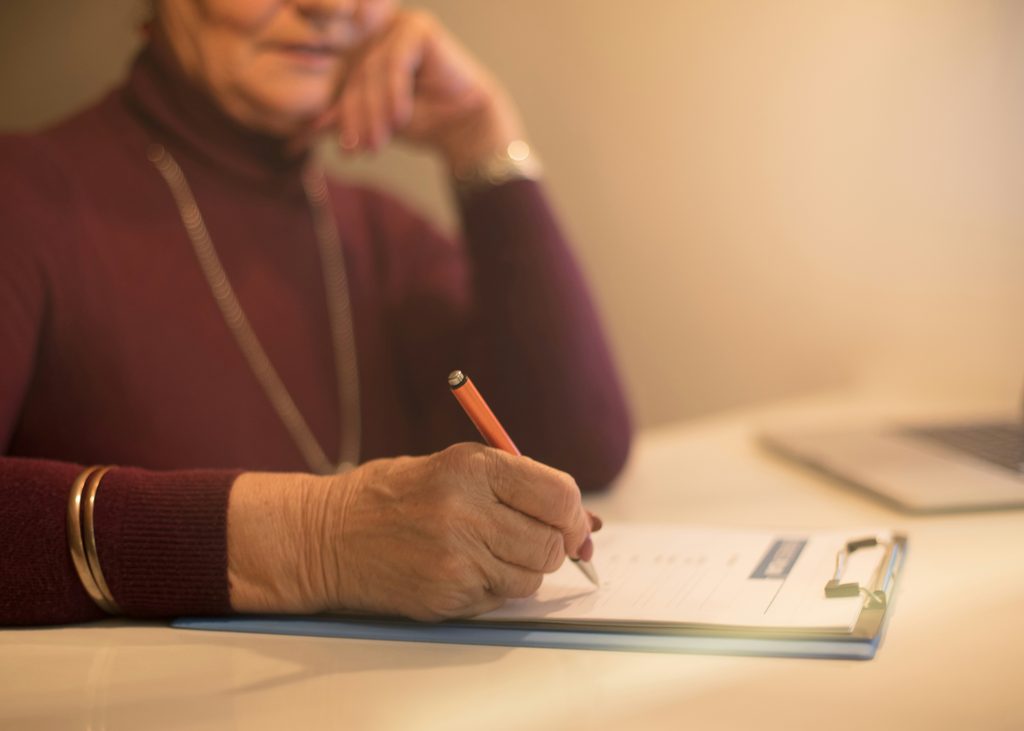 An older woman wearing a maroon top sits at a table, holding a pencil and filling out a form on a clipboard—possibly designating an attorney-in-fact in Minnesota—with a laptop nearby.