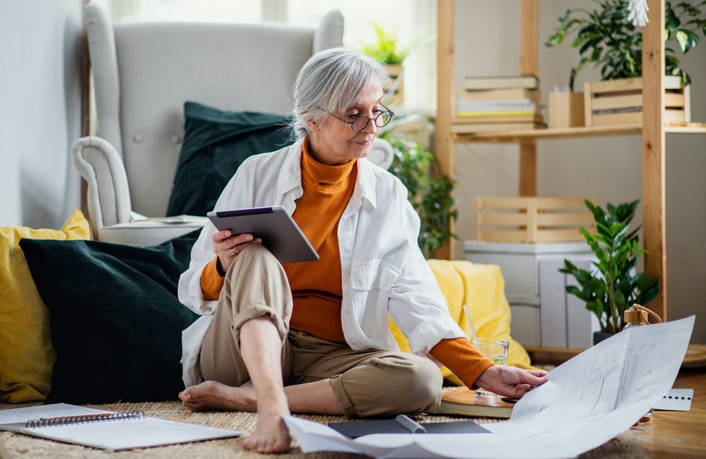 An older woman with gray hair and glasses sits on the floor, holding a tablet and examining large papers—perhaps reviewing important documents about her role as a successor trustee in Minnesota. She is surrounded by pillows, plants, and notebooks in a cozy, well-lit room.