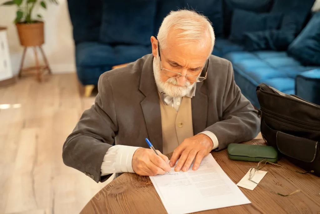 Older man in a tweed jacket signing a document at a wooden table