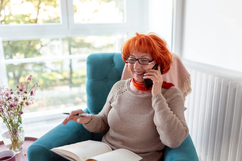 Older woman in a blue armchair smiling while she talks on the phone and takes notes