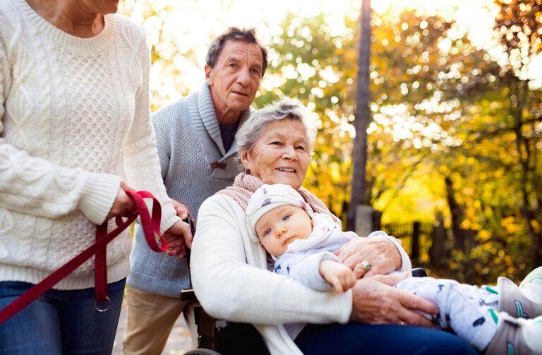 An elderly woman in a wheelchair holds a baby on her lap, while a man stands behind her and another person in a white sweater holds a dog leash. They are outdoors with trees in the background.
