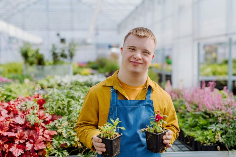 A person wearing a yellow shirt and blue apron smiles while holding two potted plants in a greenhouse filled with various colorful flowers and greenery.