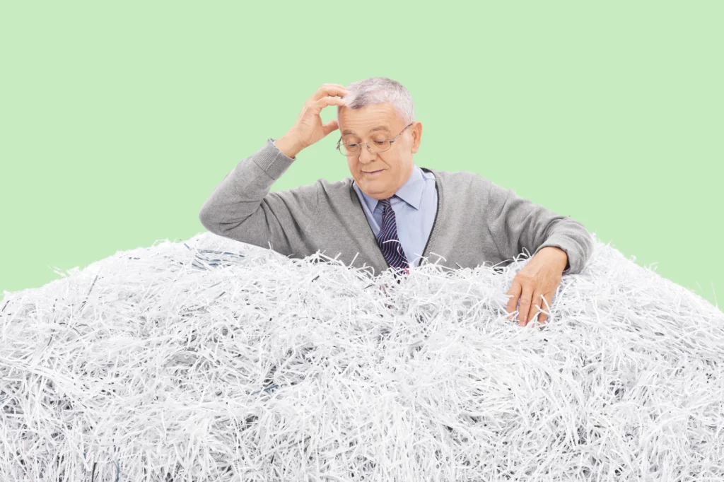 Older man in a gray cardigan scratching his head over a pile of shredded paper